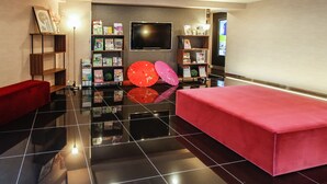 A modern lobby with a red sofa, a red bench, and a wall-mounted TV. Shelves with books and magazines are visible. - HOTEL LiVEMAX Kyoto Nijojo-kita (Kyoto)