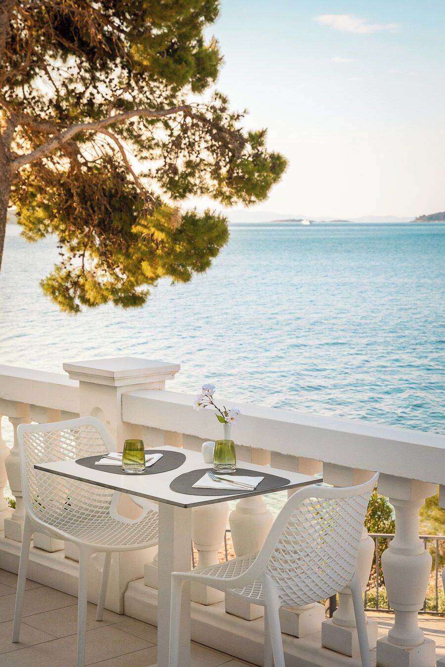 A seaside terrace with white wicker furniture, a table with a vase and glass, and a view of the sea.