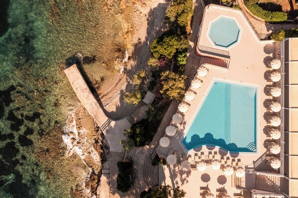 An aerial view of a resort with a large swimming pool, sun loungers, and a rocky shoreline.