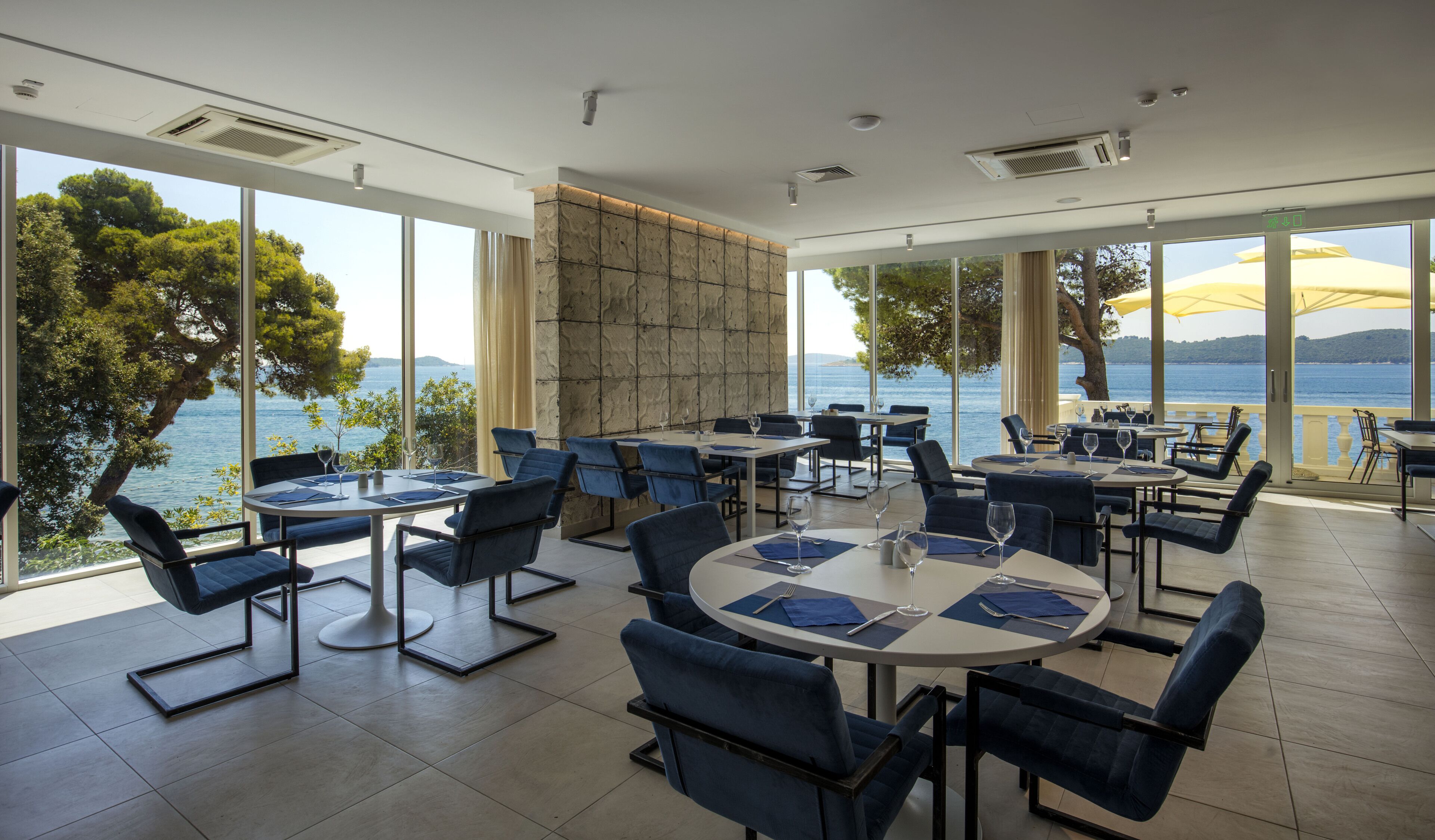 A restaurant with round tables and blue chairs, large windows with a view of the sea, and a stone wall.