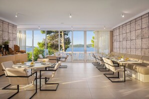 A modern dining area with tables and chairs, large windows with a view of the sea, and a stone wall. - Aminess Younique Bellevue Hotel (Orebic)