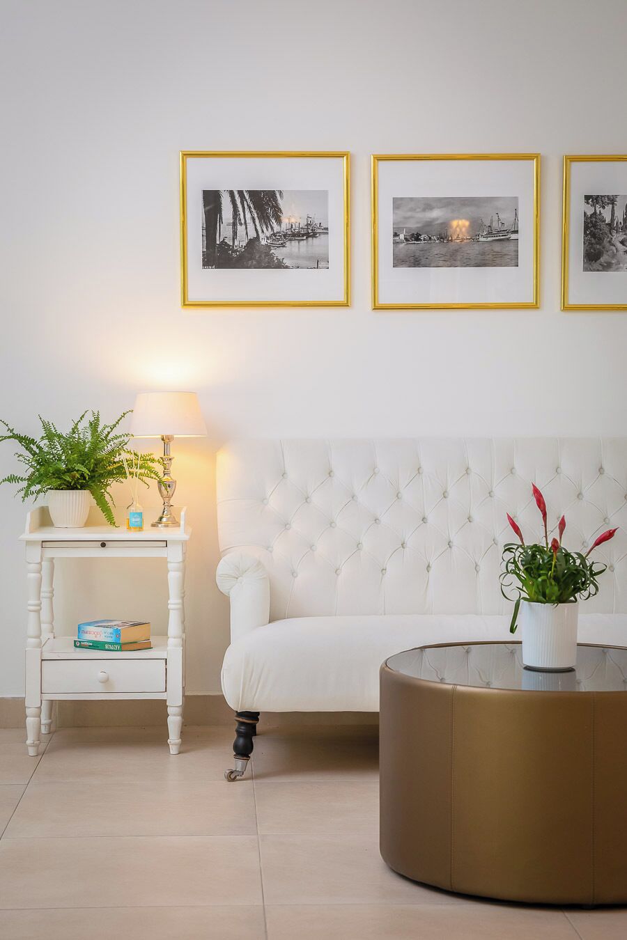 Una habitación con un sofá blanco, una mesa redonda, una mesita auxiliar blanca con una lámpara y tres cuadros en la pared.