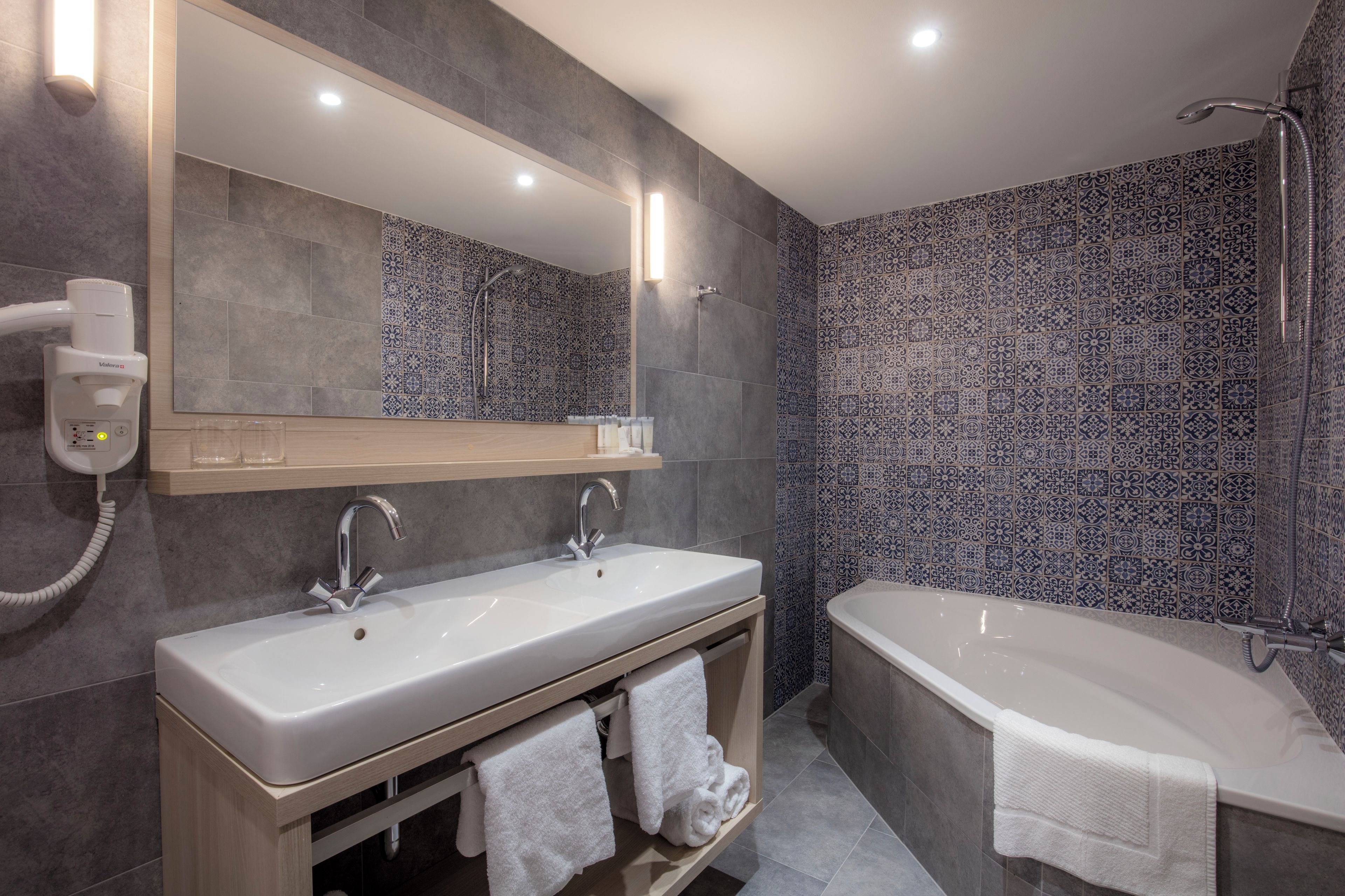 A modern bathroom with two sinks, a large mirror, a bathtub, and patterned tiles.