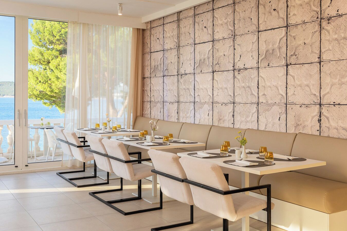 A dining area with tables set for two, a marble wall, and a view of the sea through large windows.