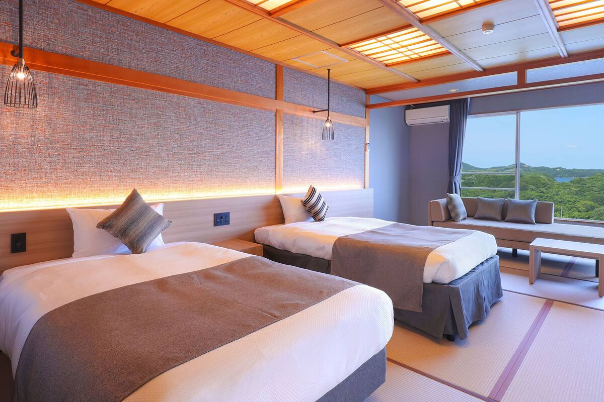A modern hotel room with two beds, a large window with a view of greenery, and a seating area with a view.