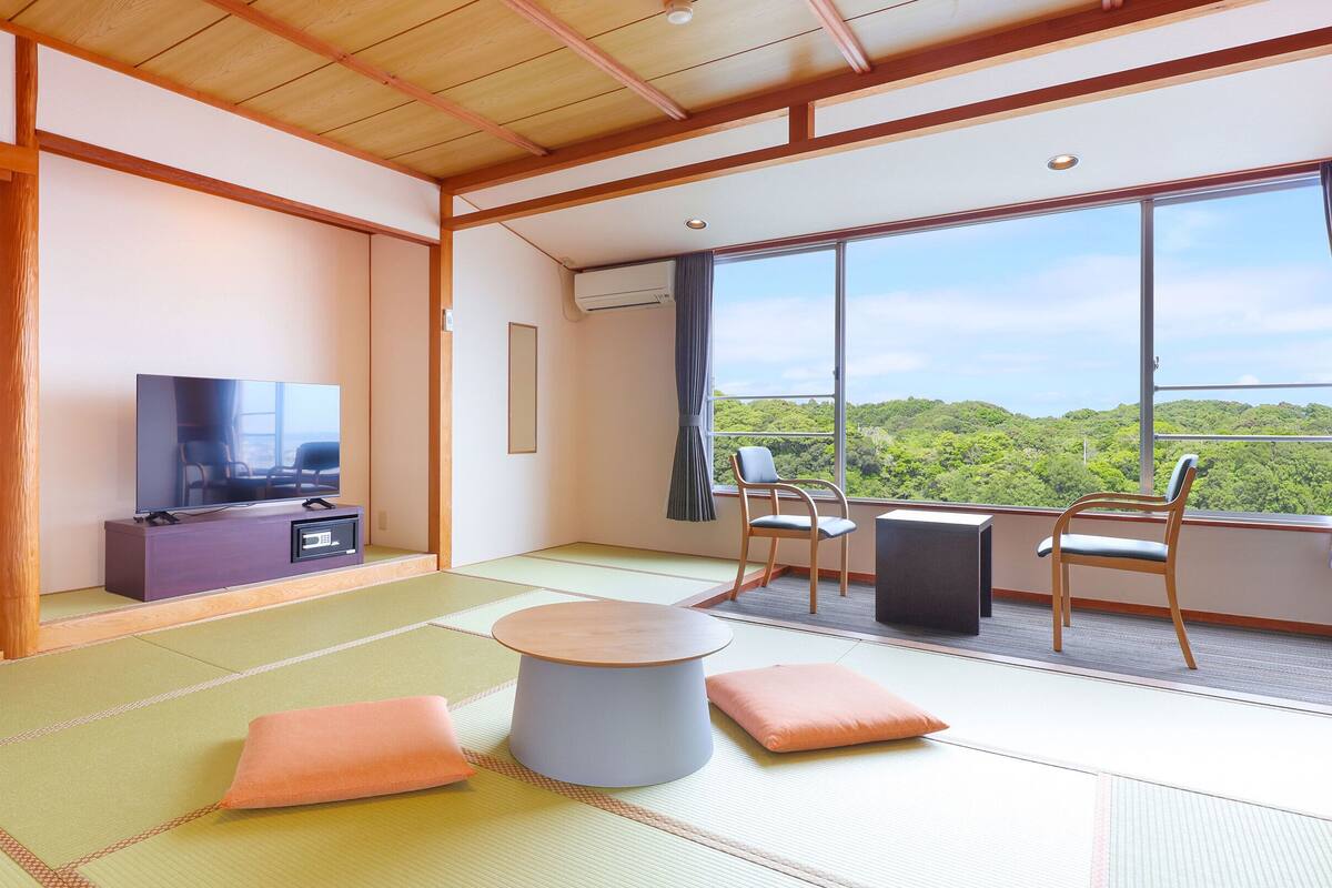 A room with tatami flooring, a television, a sofa, and a view of a forest through large windows.