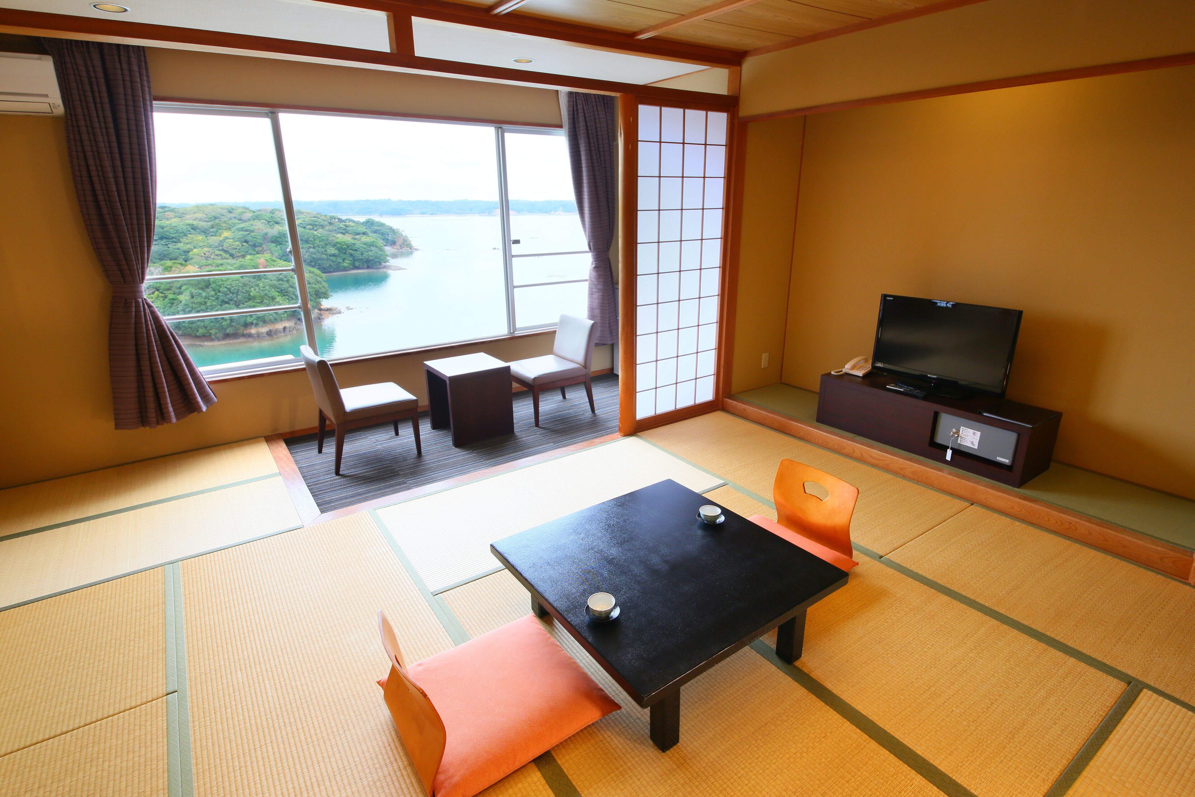 A traditional Japanese room with tatami flooring, a low table with cushions, a television, and a view of a body of water through large windows.