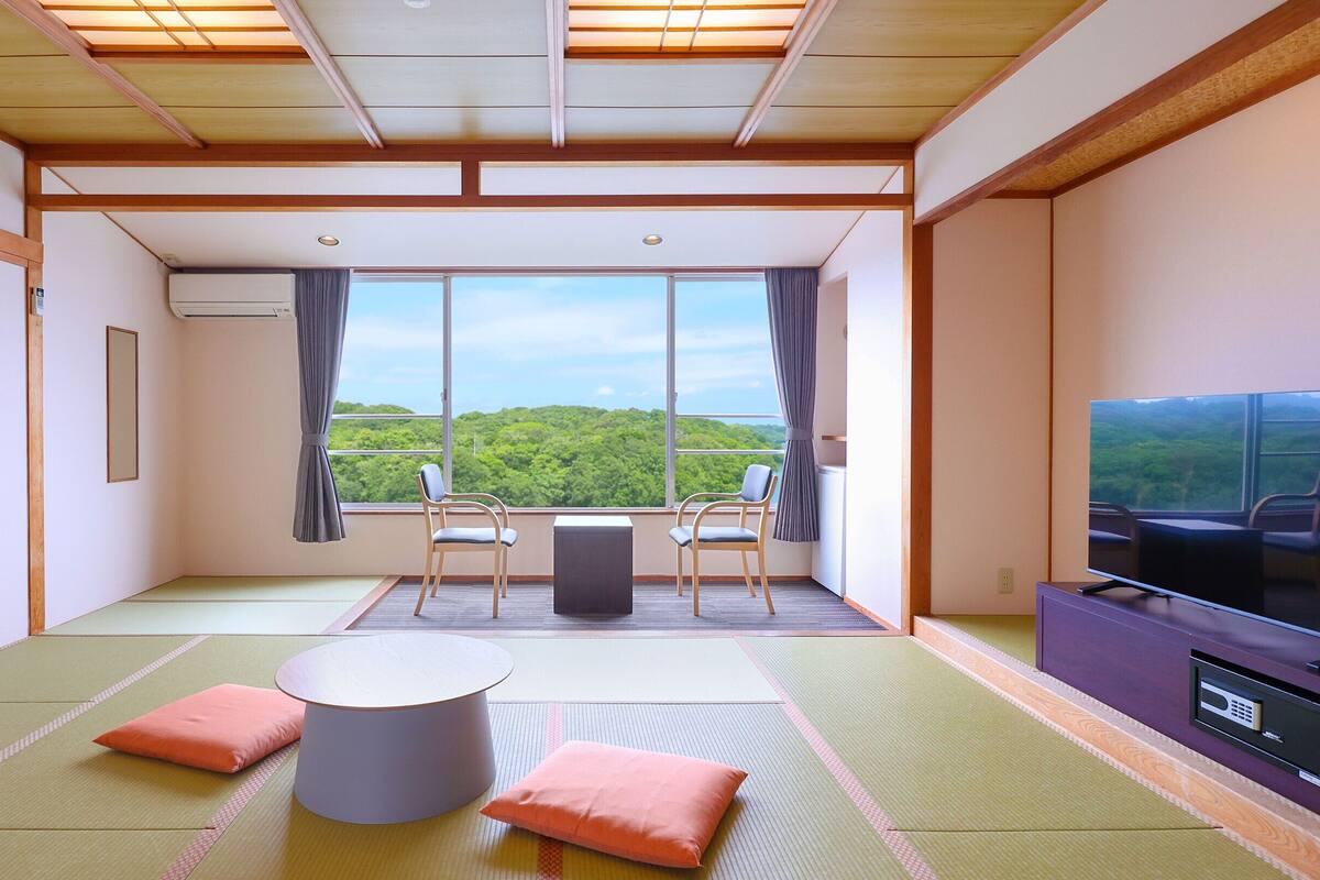 A room with tatami mats, a low table, two chairs, a TV, and a large window with a view of greenery.