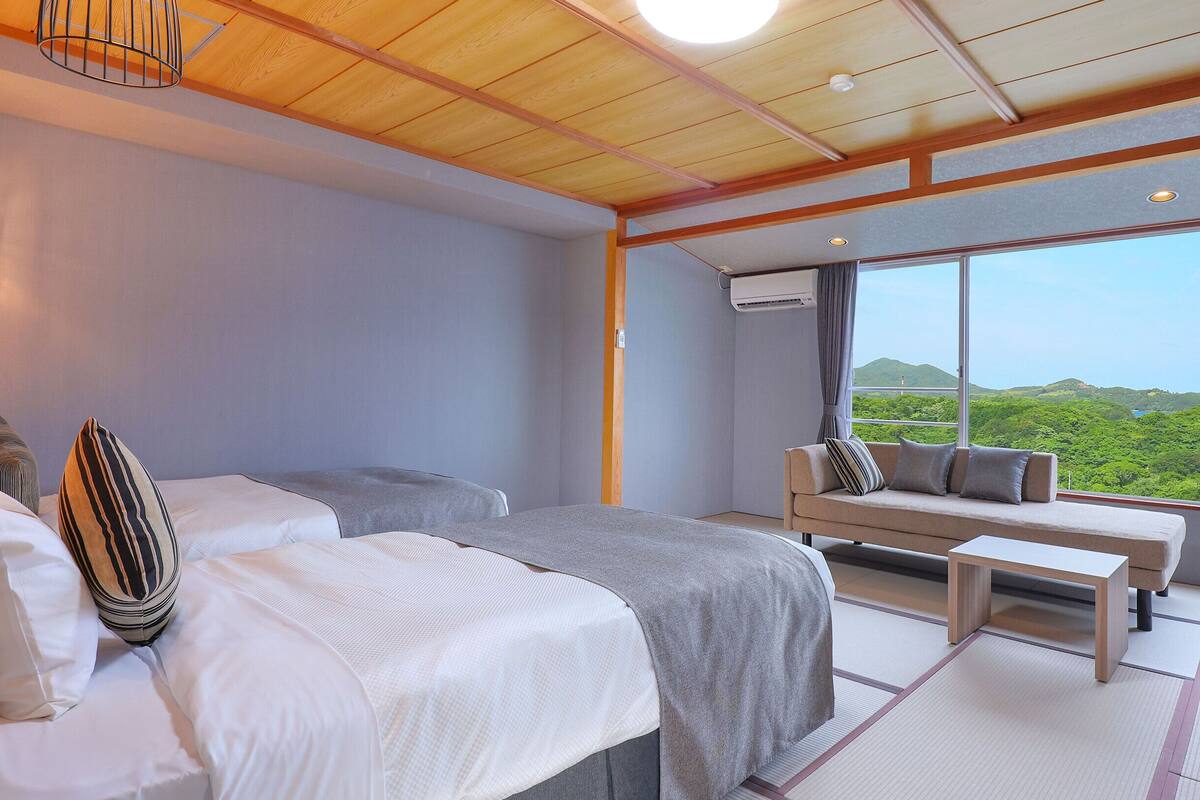 A bedroom with two beds, a sofa, a view of greenery, and a wooden ceiling.