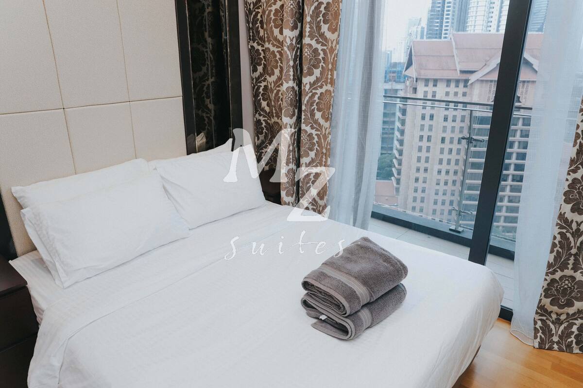 Ein Hotelzimmer mit einem großen Bett, weißer Bettwäsche, Blick auf die Stadt durch ein Fenster und einem Vorhang mit einem Blumenmuster.