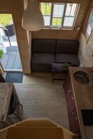 Uma cabana compacta e moderna, com interior em madeira, um sofá, uma mesa pequena, área de cozinha e vista para o exterior.