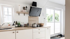 A modern kitchen with a sink, countertop, and cabinets. There is a window with blinds, a shelf with kitchen utensils, and a door with a mat. - Ferienhof Schild (Wangerland)