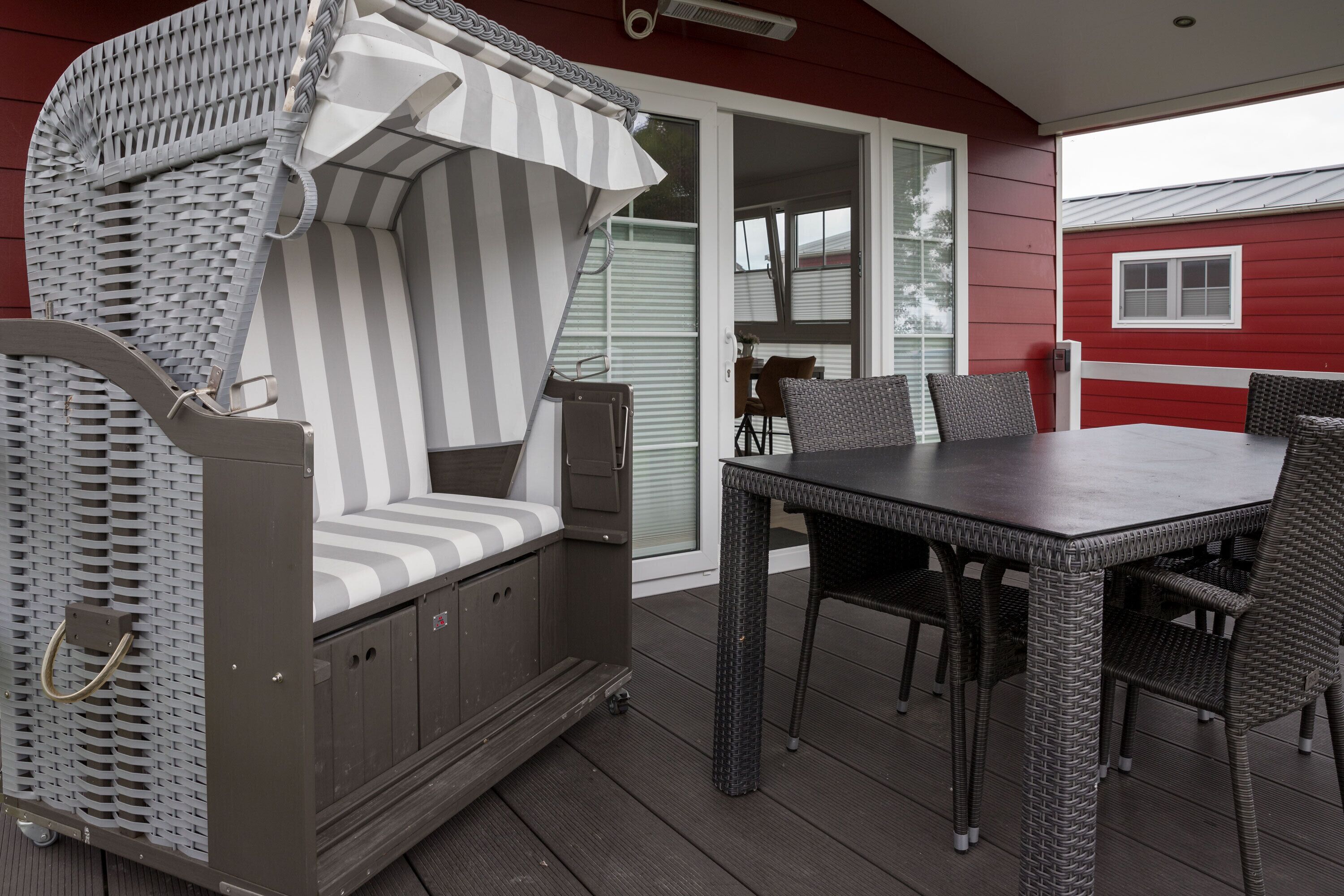 Un espace extérieur aménagé avec une zone salon couverte, comprenant une chaise en osier et une table, une porte-fenêtre coulissante, et un bâtiment rouge en arrière-plan.