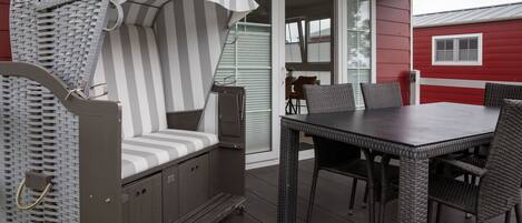 Un espace extérieur aménagé avec une zone salon couverte, comprenant une chaise en osier et une table, une porte-fenêtre coulissante, et un bâtiment rouge en arrière-plan.
