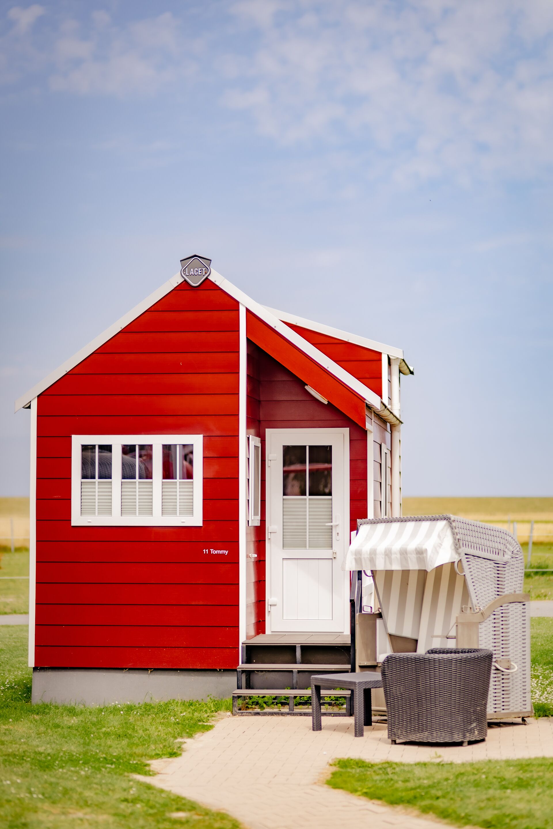 Un petit bâtiment rouge avec une porte et des fenêtres blanches, un espace extérieur abrité avec des sièges, et un chemin pavé menant à l’entrée.