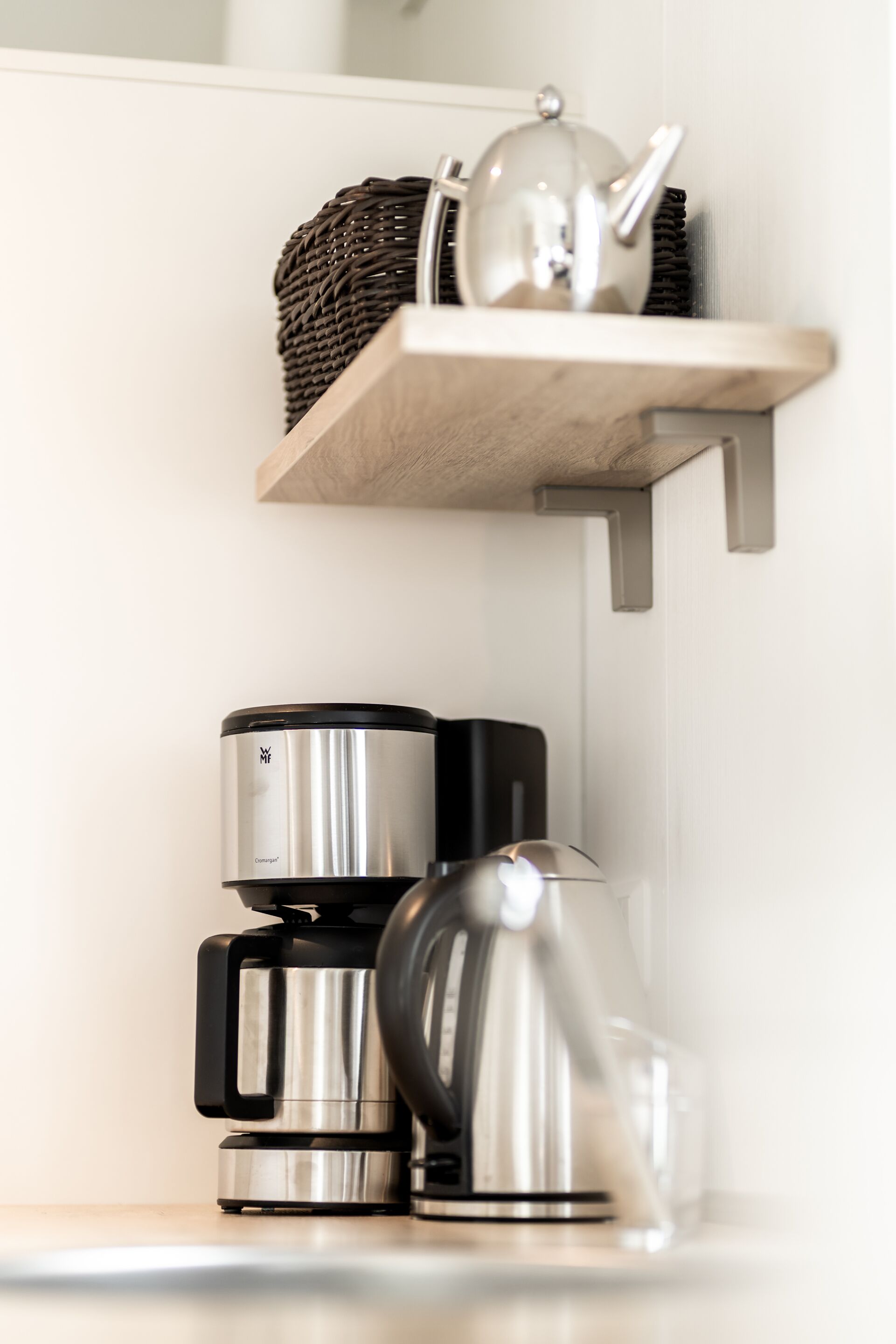 Een aanrecht met een koffiezetapparaat, een waterkoker en een mandje op een houten plank.