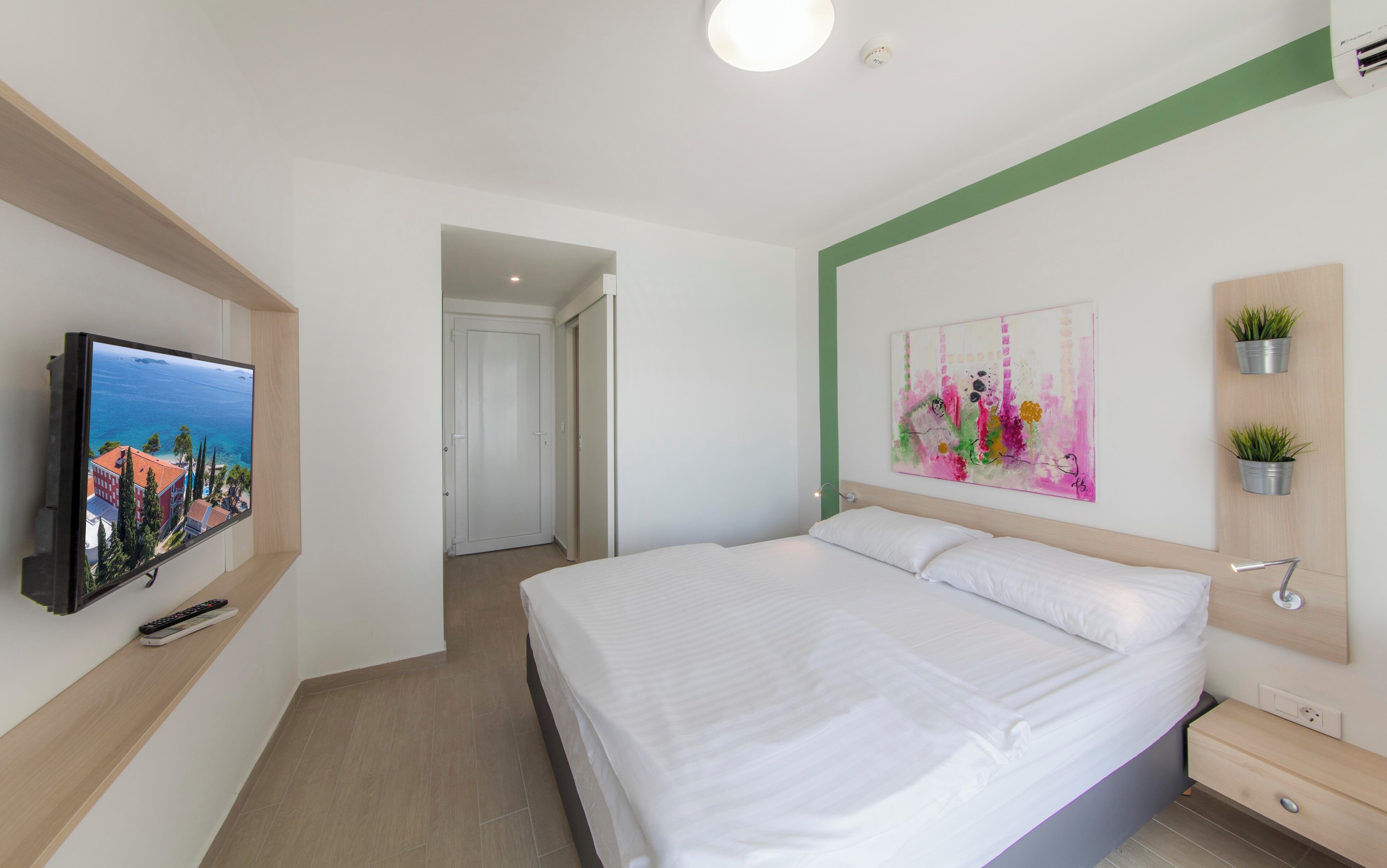 A modern hotel room with a large bed, a television mounted on the wall, a painting, and a small plant on the dresser.