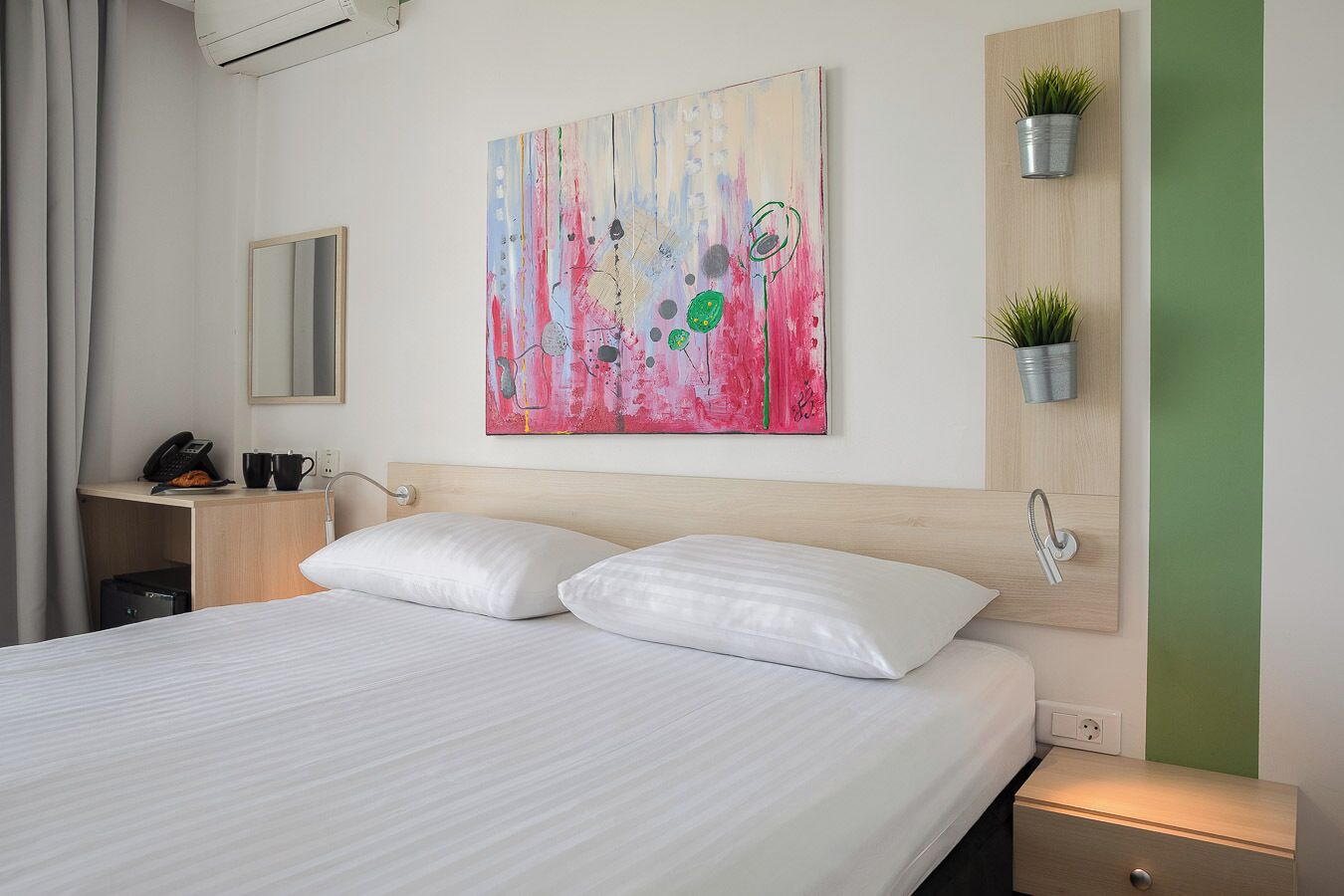 Une chambre d’hôtel comprenant un lit, une table de chevet, un bureau avec un téléphone et une tasse, un miroir fixé au mur, une peinture abstraite aux couleurs vives, et un mur vert agrémenté d’une plante en pot.