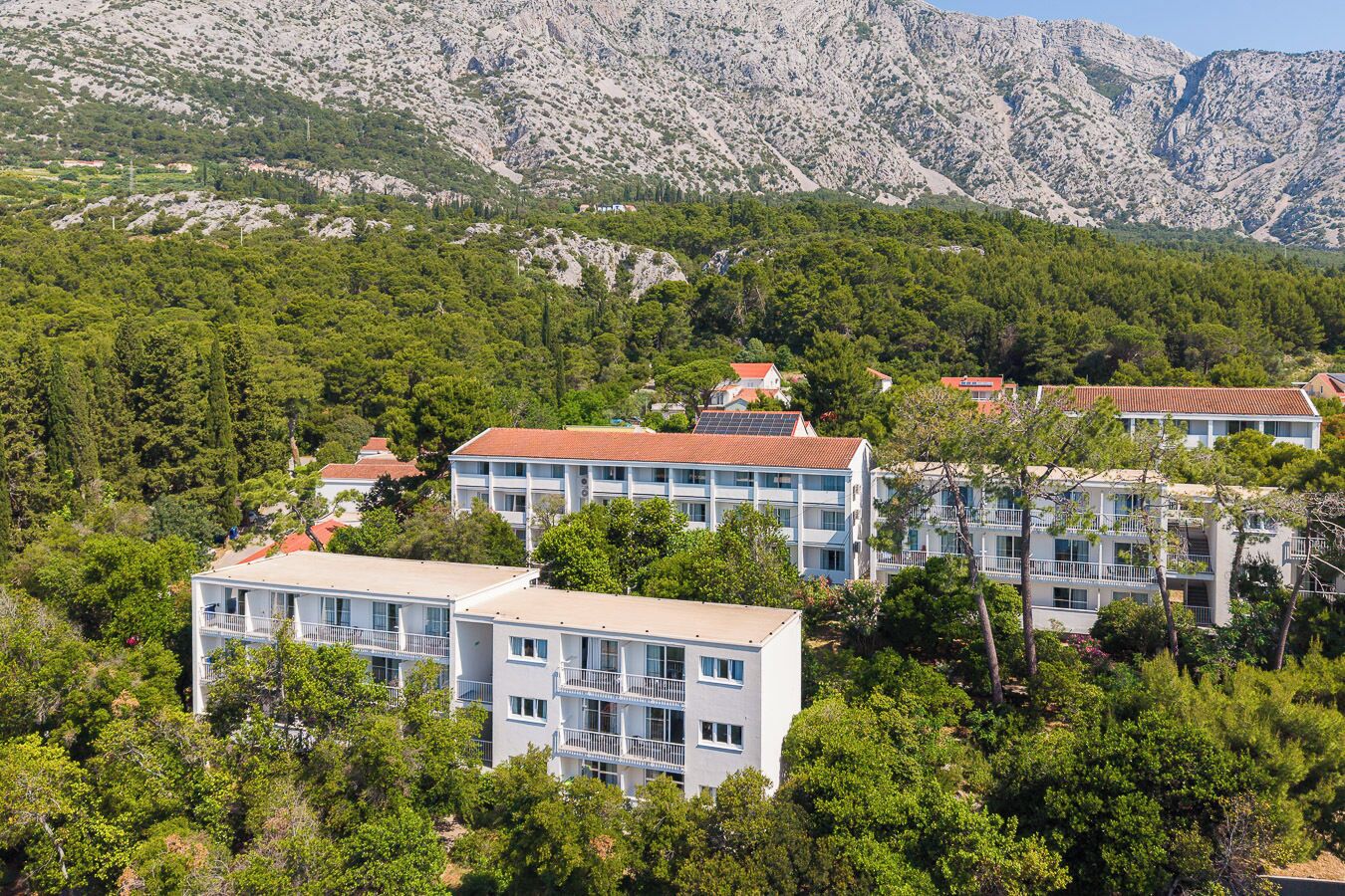 A cluster of multi-story buildings with balconies, surrounded by dense greenery and mountains in the background.
