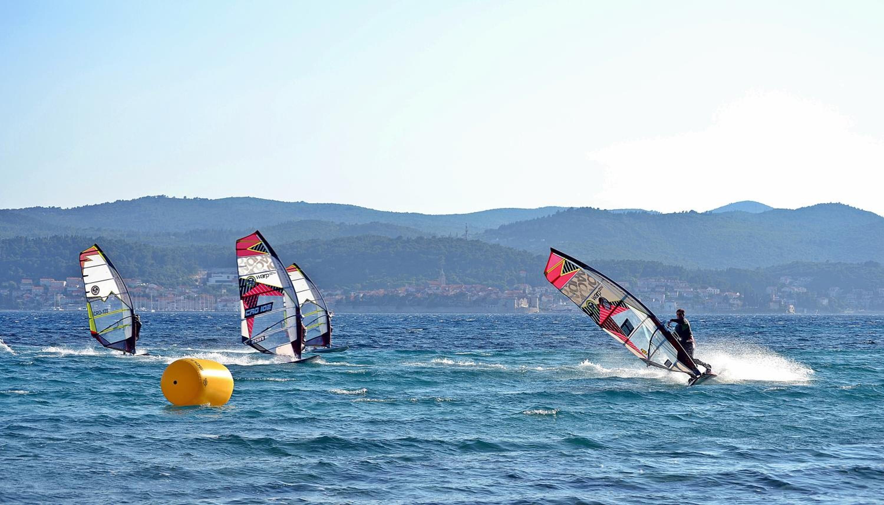 A coastal scene with three windsurfers on the water, a yellow buoy, and a mountainous backdrop.