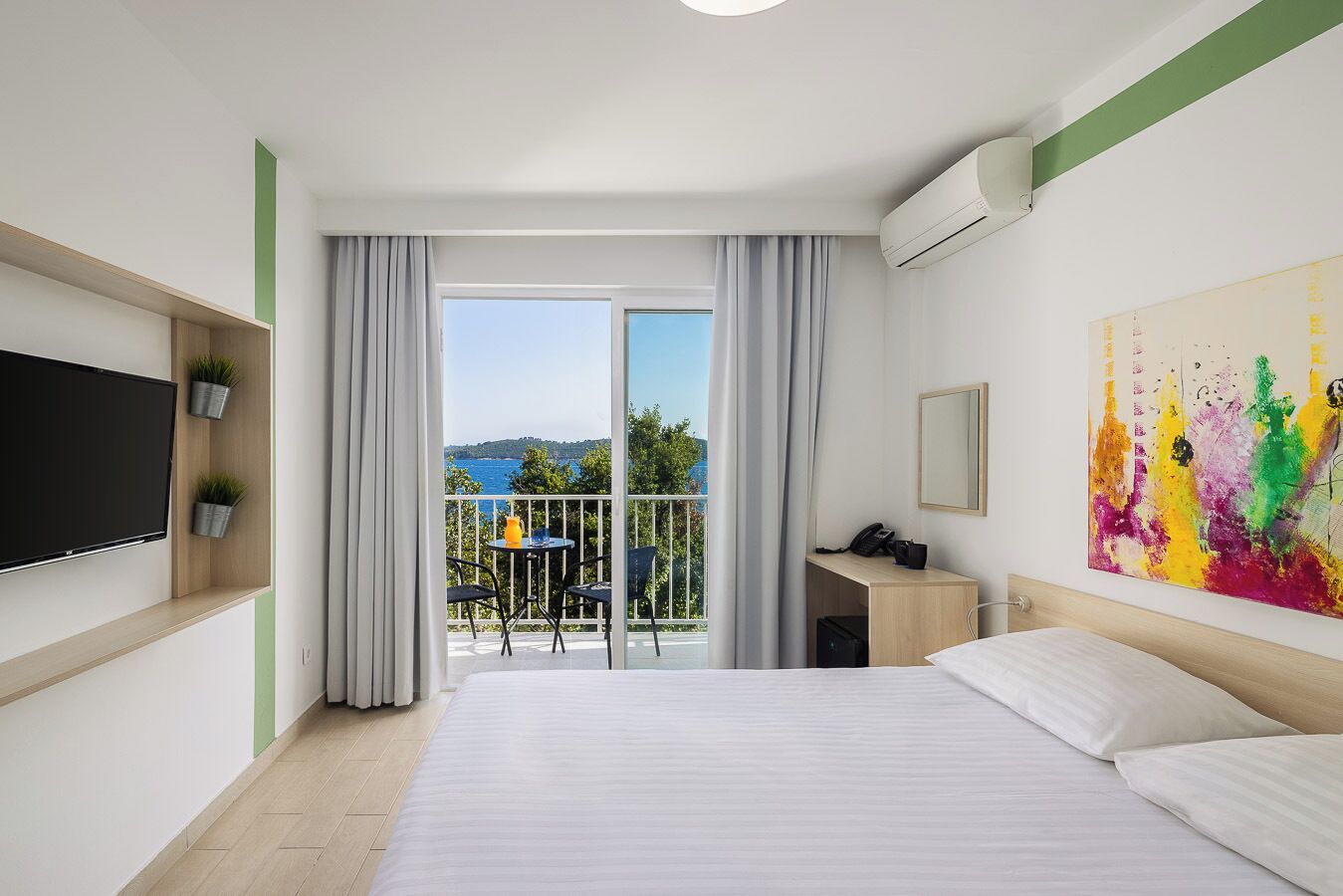 A hotel room with a bed, a TV, a desk, a chair, a balcony with a view, and a painting.