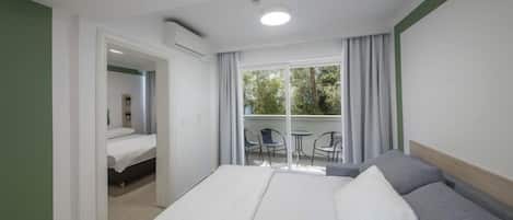 A hotel room with a bed, a balcony with chairs, and a view of trees.