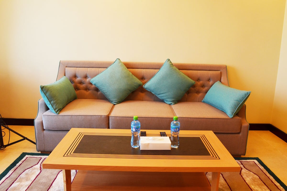 A brown tufted sofa with four teal pillows and a wooden coffee table with two water bottles and a remote control.