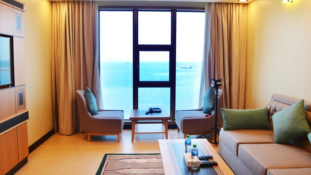 A hotel room with a large window offering a view of the sea, two armchairs, a sofa, a coffee table, and a TV on a cabinet. - Nooh Tower (Hidd)