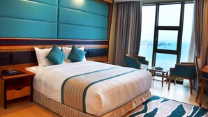 A hotel room with a large bed, a nightstand, a desk, and a view of the sea. - Nooh Tower (Hidd)