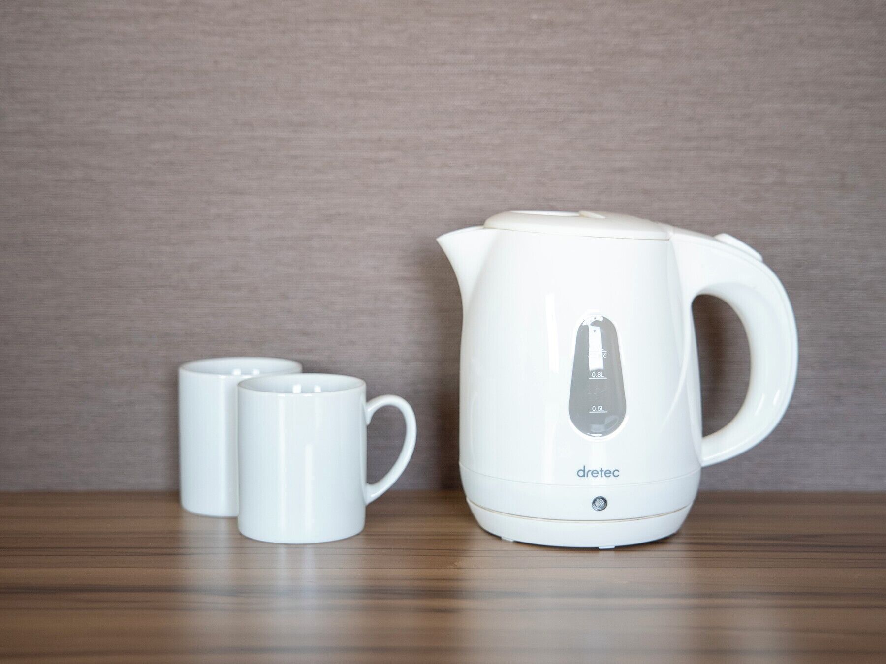 Een witte elektrische waterkoker met aanduidingen voor waterhoeveelheid en drie witte mokken op een houten oppervlak voor een gestructureerde muur.