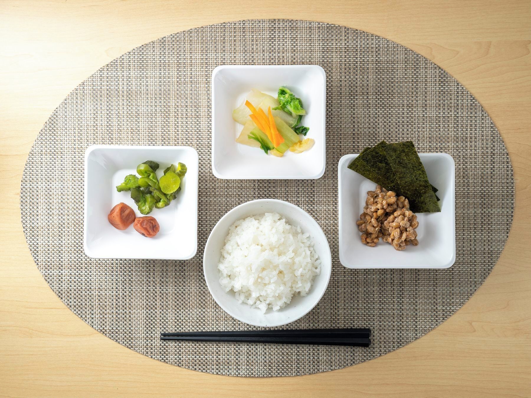 A meal setup with white bowls on a woven placemat, including rice, vegetables, and a seaweed dish.
