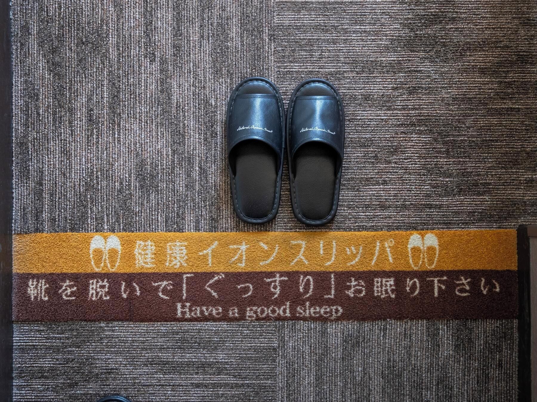 A pair of black slippers on a textured brown and gray carpet with a brown strip featuring Japanese text and English translation.