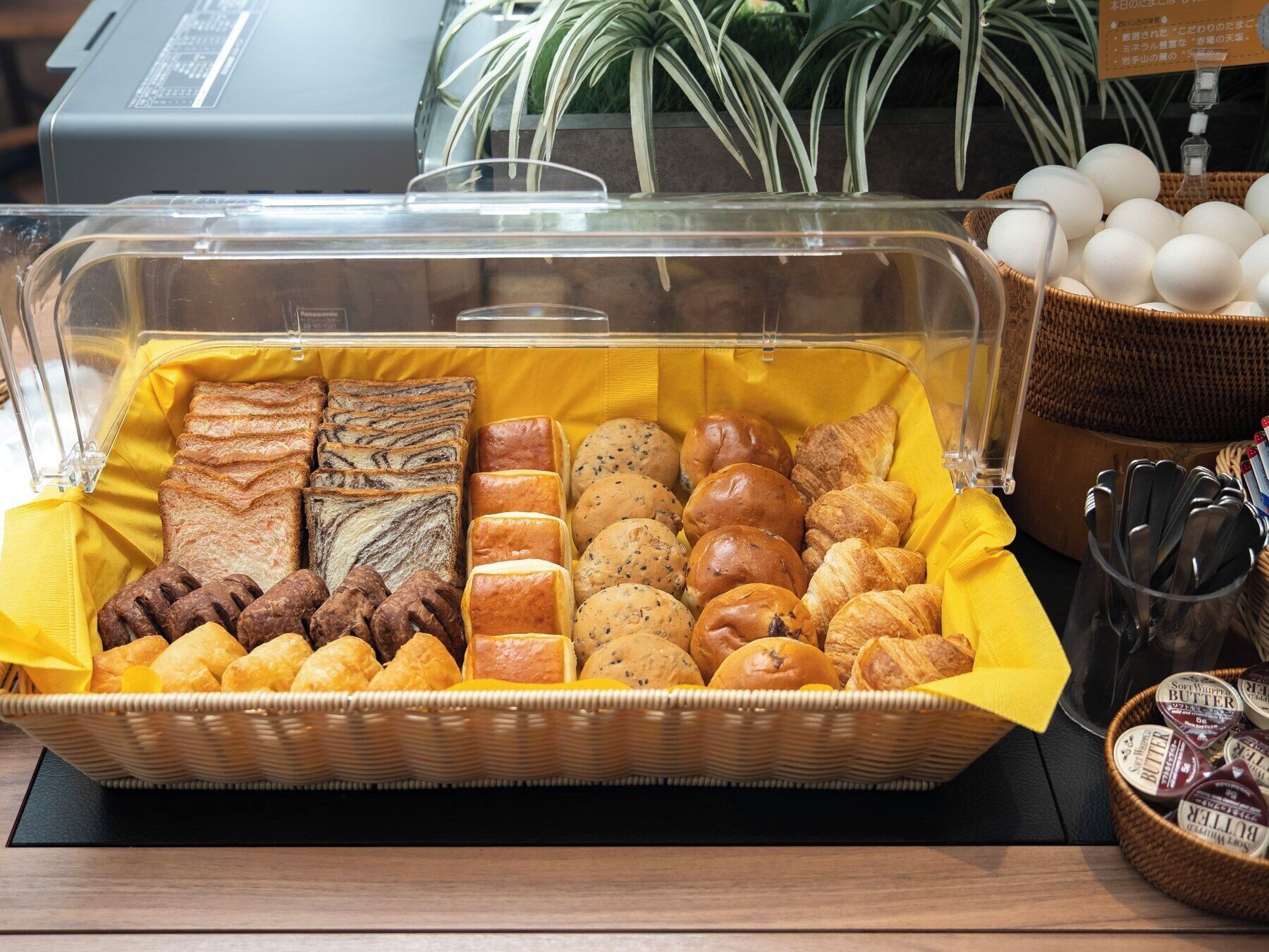 A breakfast buffet with various breads, pastries, and a basket of eggs.