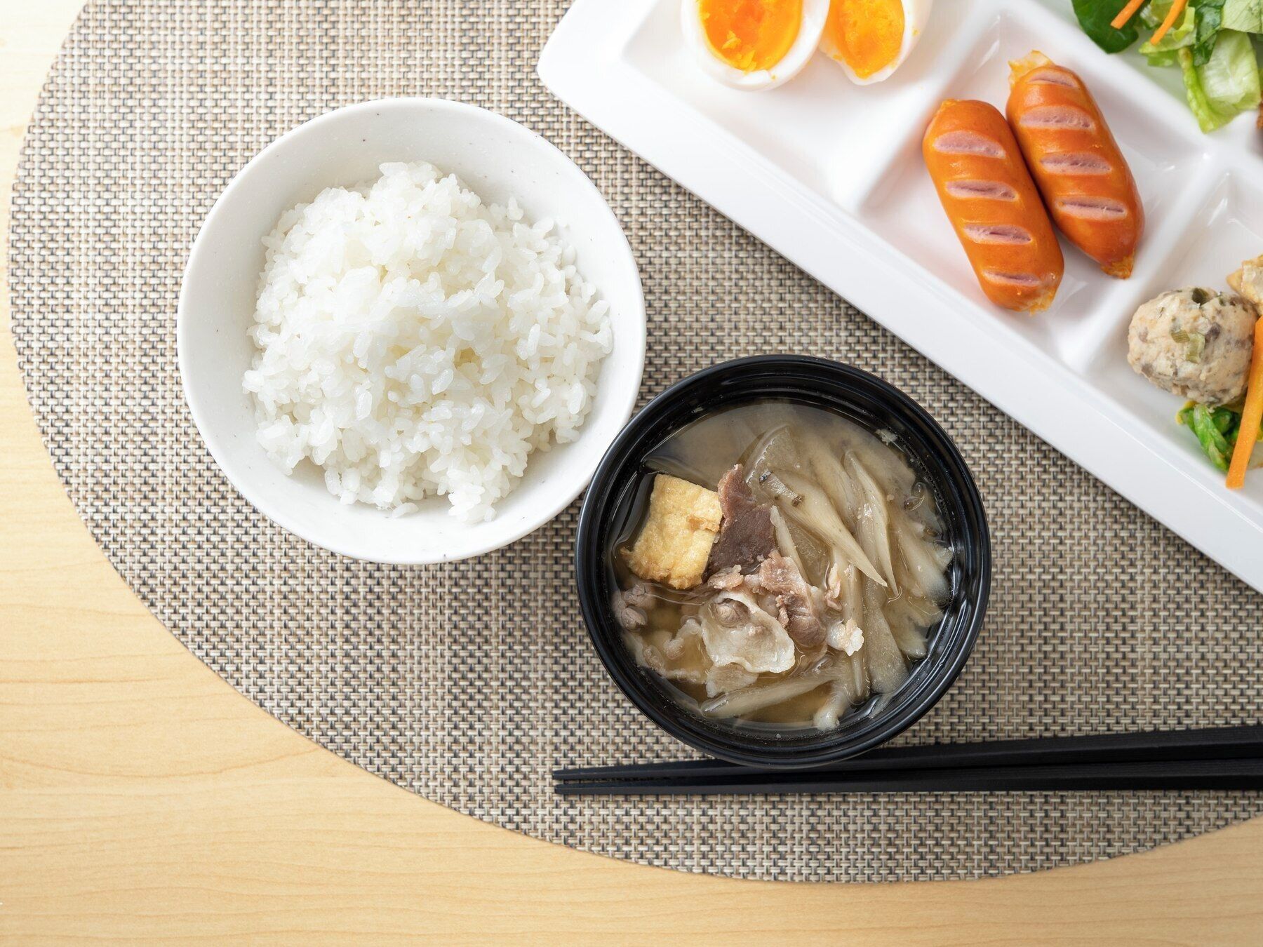 A meal consisting of white rice, a bowl of soup with meat and vegetables, and a plate with sausages and a meatball.