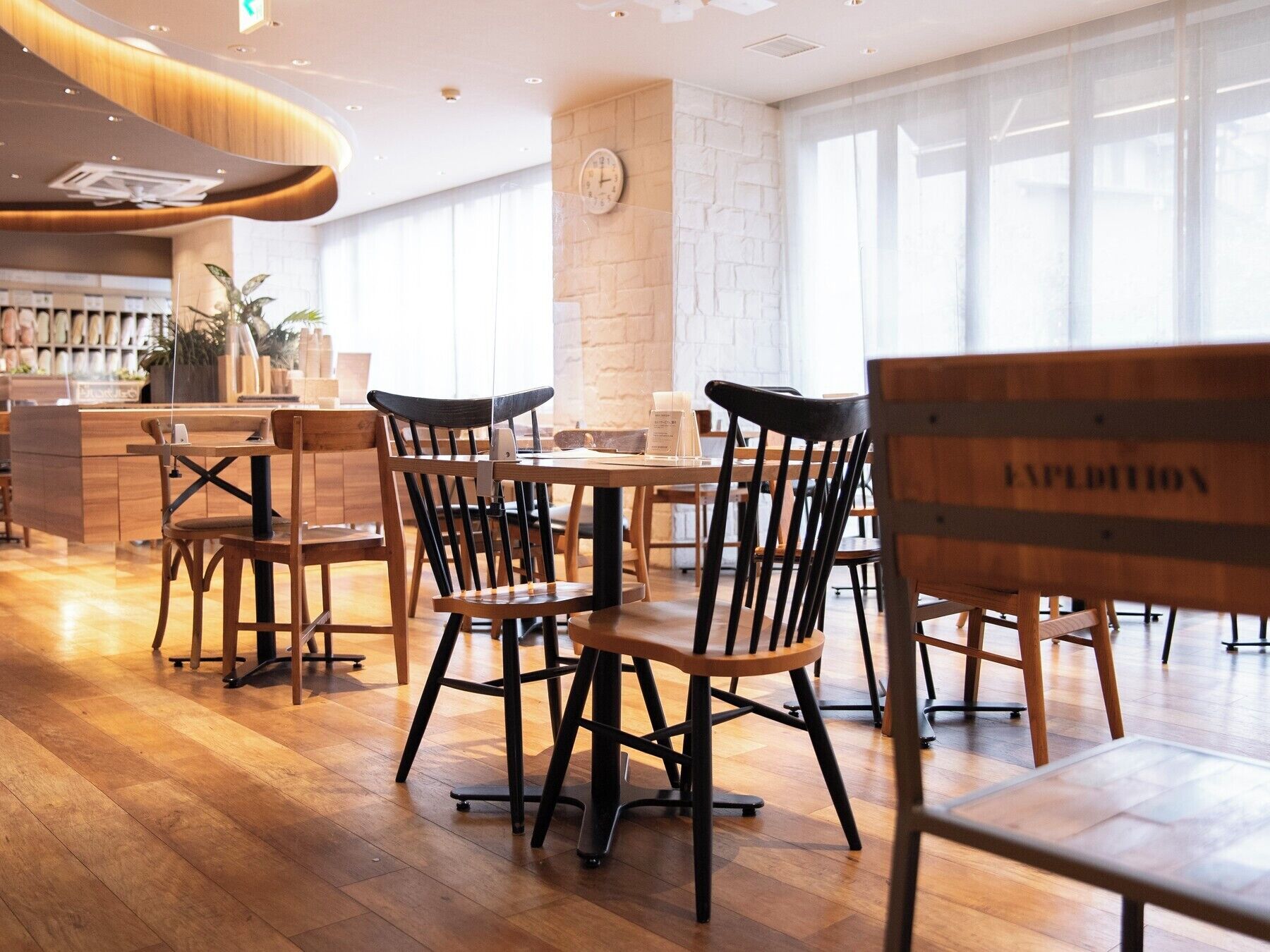 A modern restaurant interior with wooden tables and chairs, a stone wall, and large windows.