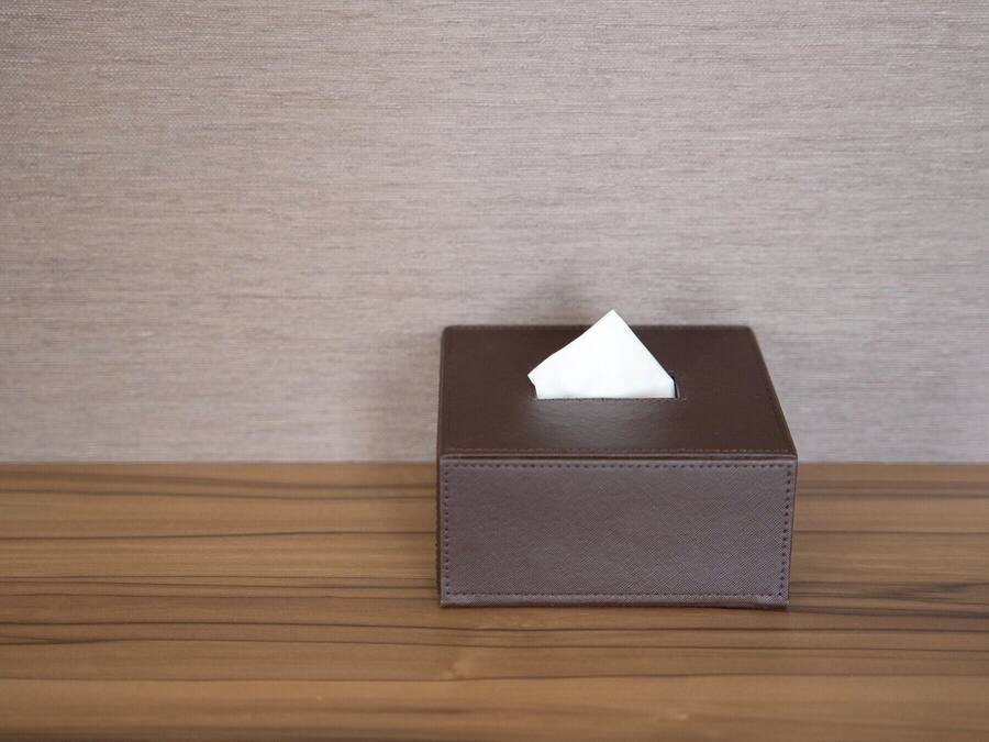 A small, dark brown, textured box with a white tissue on top, placed on a wooden surface against a plain wall.