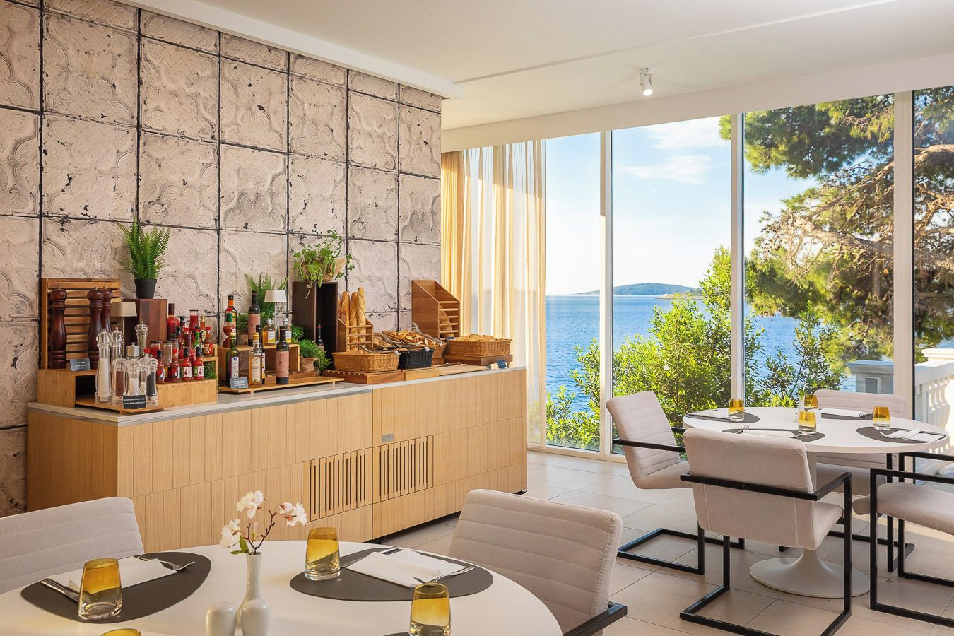 A modern dining area with a view of the sea, a wooden bar with various bottles and glasses, and a seating arrangement with tables and chairs.