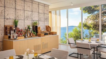 A modern dining area with a view of the sea, a wooden bar with various bottles and glasses, and a seating arrangement with tables and chairs.