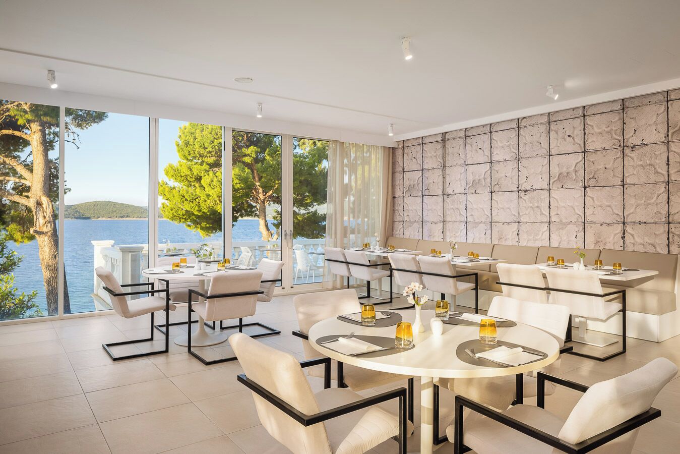A modern dining area with multiple tables and chairs, large windows with a view of the sea, and a textured wall.