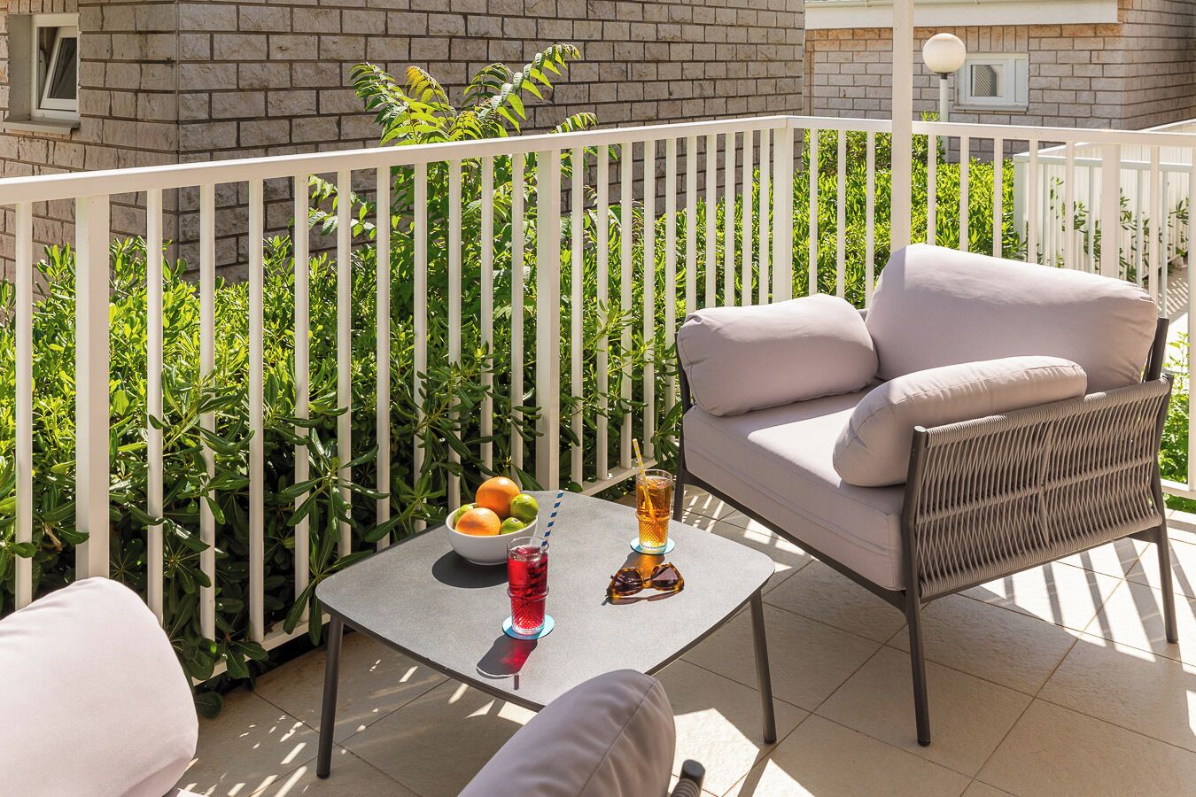 A balcony with a white railing, a wicker sofa, a small table with a bowl of fruit and a drink, and a tiled floor.