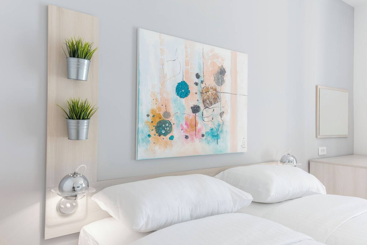 Una habitación de hotel moderna con una cama, dos macetas colgantes con plantas verdes, un cuadro abstracto colorido y una mesita de noche con una lámpara.