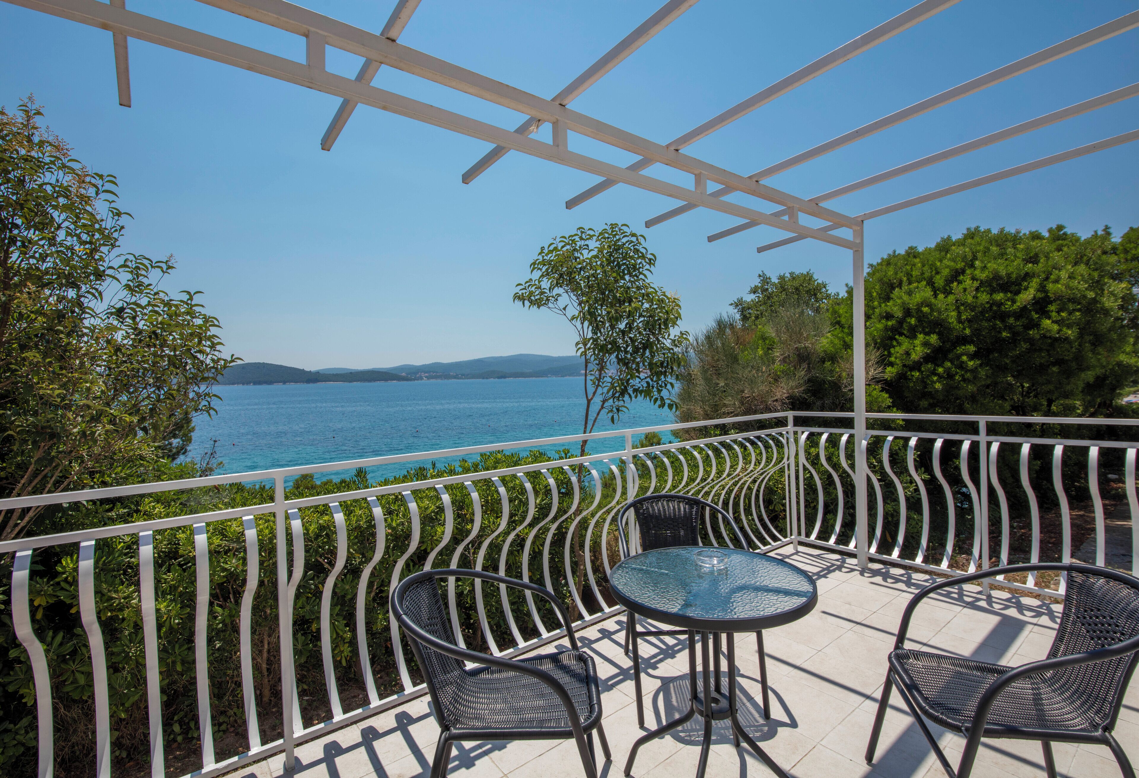 Un balcon doté d’une balustrade blanche, d’une table en verre et de quatre chaises, offrant une vue sur une étendue d’eau et sur la verdure.
