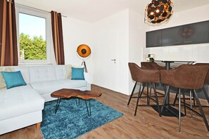 Ein modernes Wohnzimmer mit einem weißen Sofa, einem Holztisch und einem blauen Teppich. Es gibt einen Essbereich mit Tisch und Stühlen sowie eine Küche mit dunklen Schränken.