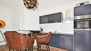 Moderni keittiö, jossa on tummat kaapit, ruostumattomasta teräksestä valmistettu liesi ja sisäänrakennettu uuni. Keittiössä on myös ruokailutila, jossa on kaksi ruskeaa tuolia ja lasipöytä.