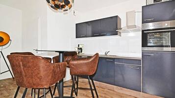 A modern kitchen with dark cabinets, a stainless steel range, and a built-in oven. There is a dining area with two brown chairs and a glass table.