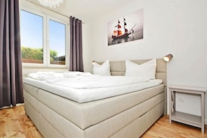 Cama bem arrumada com lençóis brancos em quarto com janela ampla, pintura de um navio na parede e mesa de cabeceira com abajur.