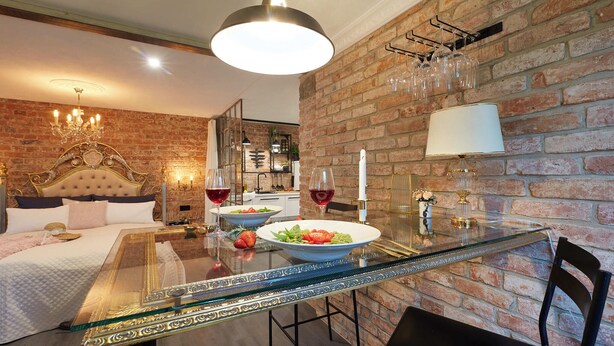 Habitación de hotel con pared de ladrillo, una mesa de comedor de vidrio, cama con cabecera dorada y candelabro.