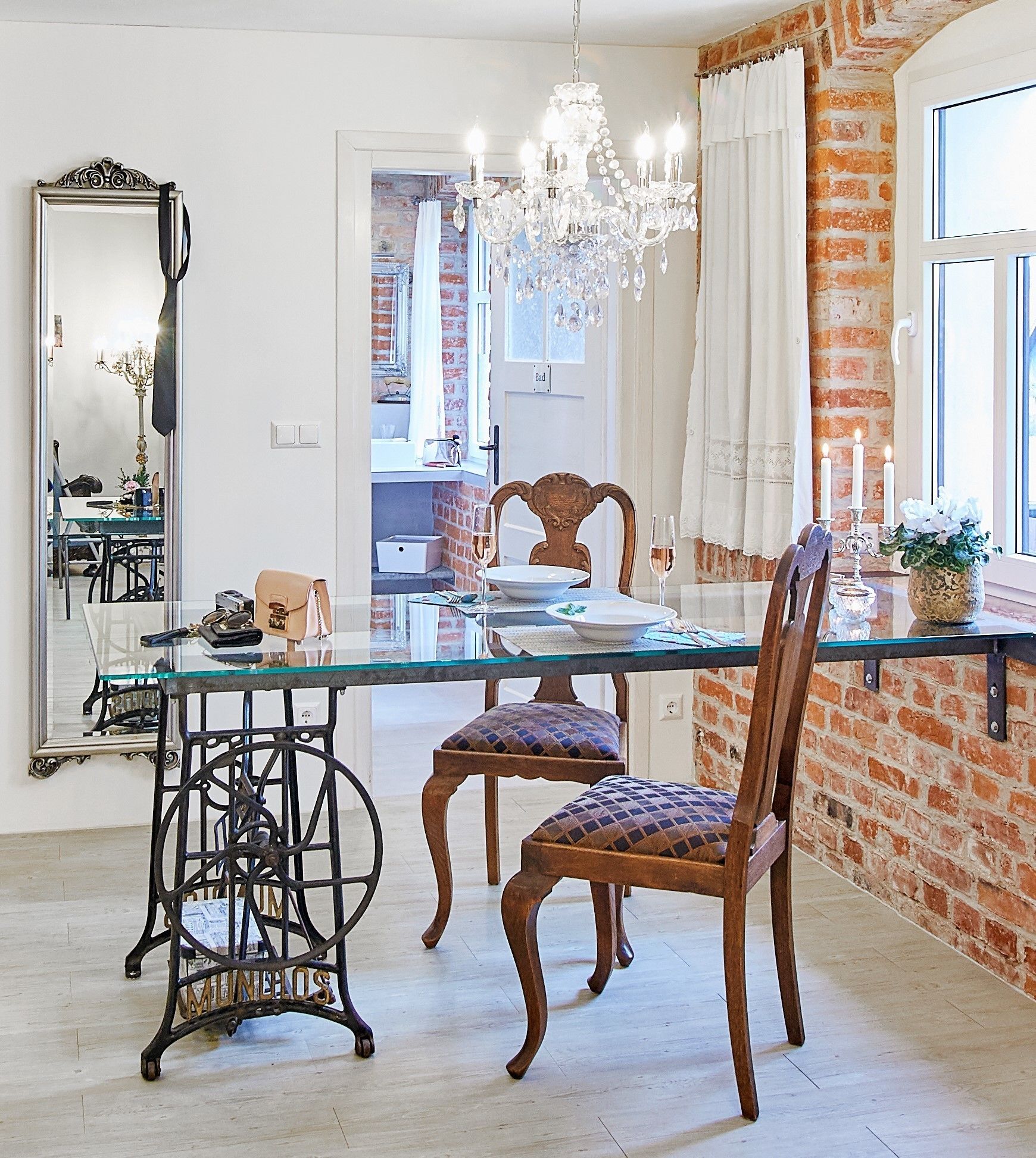 A dining area with a glass table, two wooden chairs, a chandelier, and a brick wall.