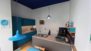 Una cocina compacta con armarios color turquesa, una puerta blanca, una lámpara colgante azul y un pequeño comedor con mesa y silla de madera.
