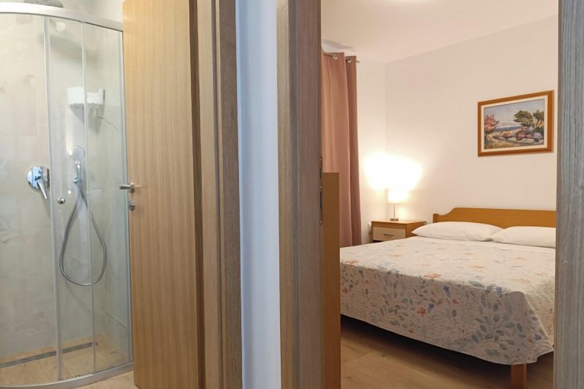 A bedroom with a bed, a nightstand, a lamp, a painting on the wall, and a shower area.